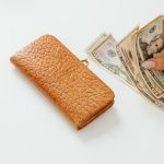 A hand counting American dollar bills next to a brown leather wallet on a white surface.